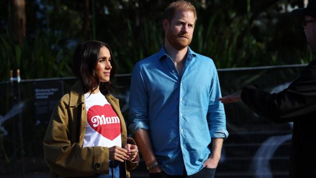 Meghan Markle und Prinz Harry stehen im Freien und unterhalten sich mit einer Person, die nur teilweise im Vordergrund zu sehen ist.