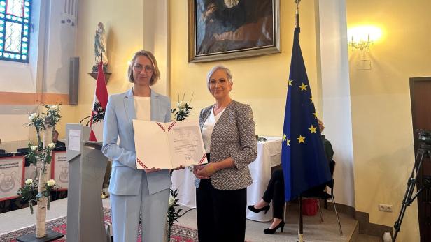 Zwei Frauen halten eine Urkunde vor Flaggen Österreichs und der EU in einem Raum.