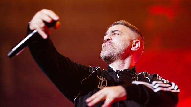 Bushido mit Bart und schwarzer Trainingsjacke hält ein Mikrofon und gestikuliert energisch vor rotem Hintergrund.