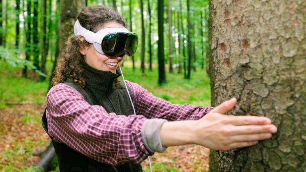 Eine Frau mit VR-Brille umarmt lachend einen Baum im Wald.