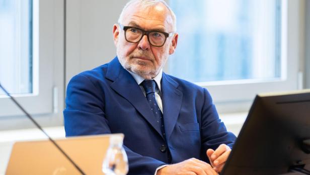 Ein älterer Mann im blauen Anzug sitzt an einem Tisch mit Laptop und Monitor.