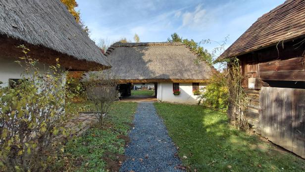 Ein schmaler Weg führt zu einem weißen Haus mit Strohdach, umgeben von alten Holzhäusern und herbstlicher Vegetation.