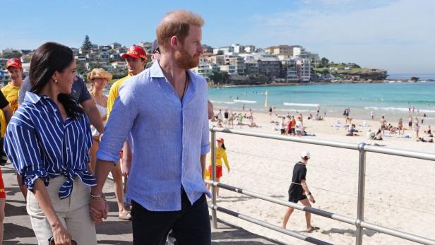 Harry und Meghan spazieren Hand in Hand am Bondi Beach entlang, im Hintergrund das Meer und Badegäste.