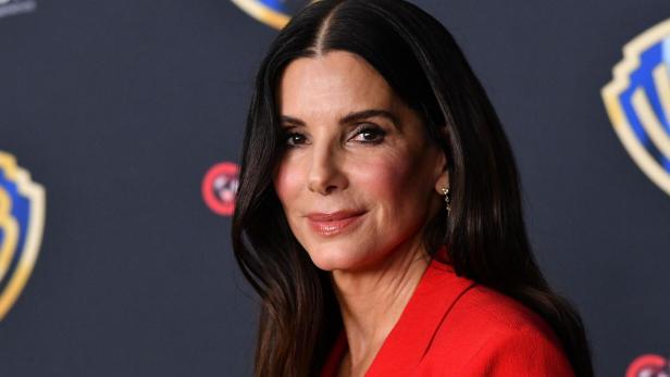 Sandra Bullock trägt einen roten Blazer vor einem dunklen Hintergrund mit dem Warner Bros. Logo.