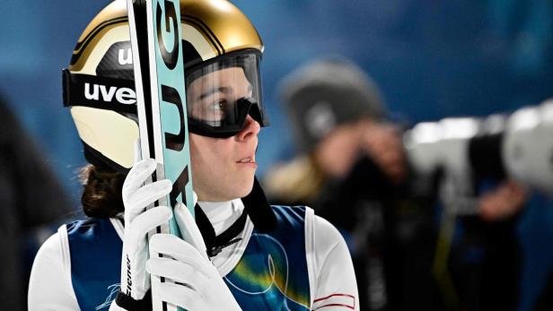 Eine Skispringerin mit goldenem Helm und Skibrille hält ihre Ski fest.