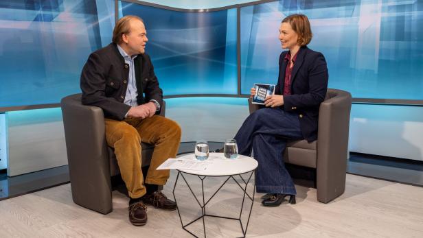 Zwei Personen sitzen sich in einem modernen Studio gegenüber, zwischen ihnen steht ein kleiner Tisch mit zwei Gläsern Wasser.
