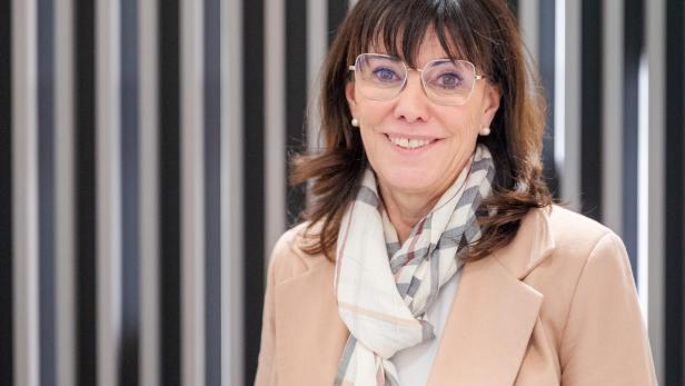 Eine lächelnde Frau mit Brille und hellem Mantel steht vor einem gestreiften Hintergrund.