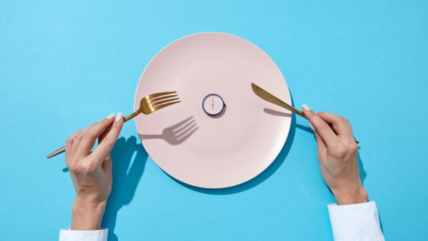 White plate with round whatch shows six o'clock served knife and fork in a girl's hands on a blue background. Time to eat and diet concept. Top view.
