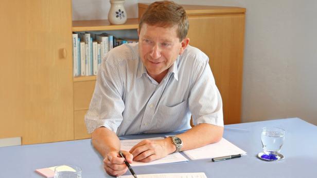 Mann sitzt an einem Tisch mit Unterlagen und Glas Wasser, Bücherregal im Hintergrund.