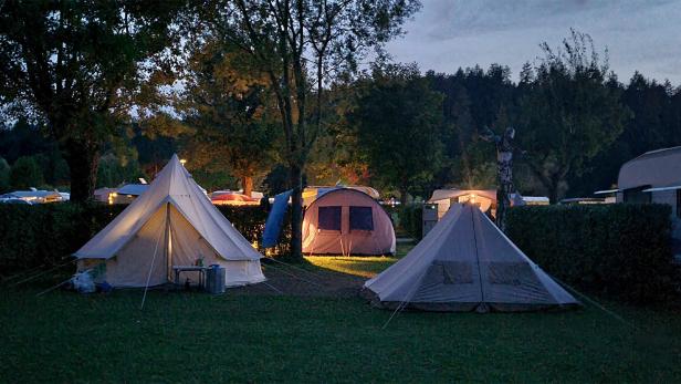 Zelte und Wohnwagen stehen abends beleuchtet auf einer grünen Wiese zwischen Bäumen.
