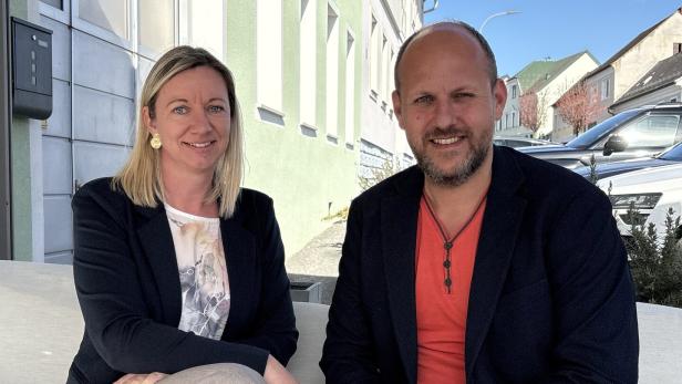 Zwei Personen sitzen nebeneinander auf einem Sofa im Freien und lächeln in die Kamera.