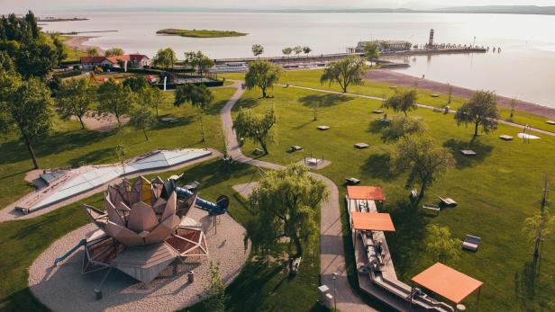 Großer grüner Park mit Spielplatz, Wegen, Bäumen und Sitzbereichen am Ufer eines ruhigen Sees mit Steg und Leuchtturm.