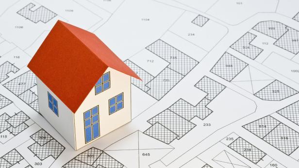 Ein Modellhaus mit rotem Dach steht auf einem detaillierten Stadtplan mit Grundstücksparzellen und Hausnummern.