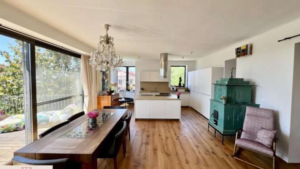 Heller, offener Wohn- und Essbereich mit Holzboden, großem Esstisch, Kronleuchter, moderner Küche und grünem Kachelofen.