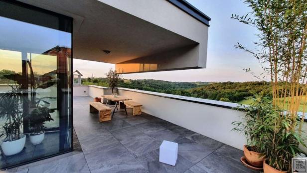 Große Terrasse mit Holztisch, Bänken, Pflanzen in Töpfen und weitem Blick auf grüne Hügel und Wälder.