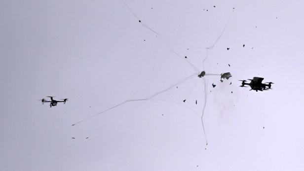 Zwei Drohnen schweben vor hellem Himmel, während eine Explosion oder Beschädigung Trümmer und Rauchspuren erzeugt.