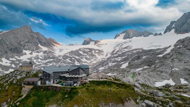 Berghütte mit mehreren Gebäuden auf grünem Hang, umgeben von schneebedeckten Felsen und hohen Bergen unter bewölktem Himmel.