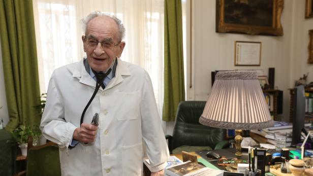 Ein lächelnder älterer Arzt mit Stethoskop und weißem Kittel steht in einem Büro.