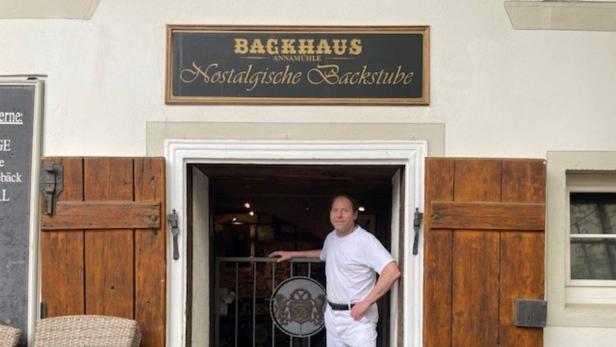 Ein Mann in weißer Kleidung steht in der Tür einer traditionellen Bäckerei mit Holzläden und nostalgischem Schild.
