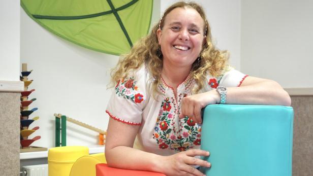 Eine lachende Frau mit lockigem Haar lehnt an bunten Schaumstoffblöcken in einem freundlich eingerichteten Raum.