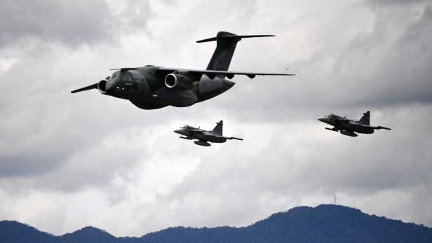 Drei Militärflugzeuge fliegen in Formation vor bewölktem Himmel über einer bewaldeten Berglandschaft.