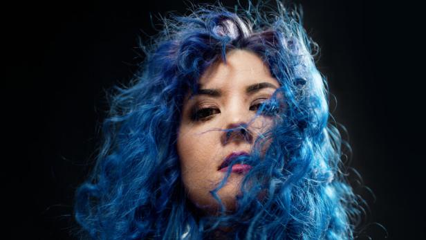 Paenda steht mit ihren blau gefärbten, lockigen Haaren und hellem Oberteil vor einem schwarzen Hintergrund.