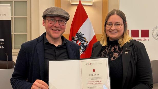Zwei Personen stehen lächelnd nebeneinander und halten gemeinsam eine Urkunde vor einer österreichischen Flagge.