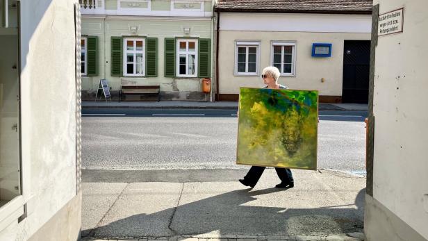 Eine Person mit heller Kurzhaarfrisur trägt ein großes, buntes Gemälde über eine sonnige Straße vor Altbauten.