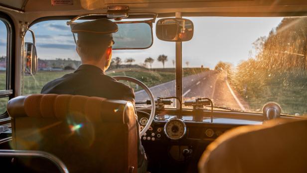 Busfahrer steuert bei Sonnenuntergang einen Oldtimerbus über eine Landstraße.