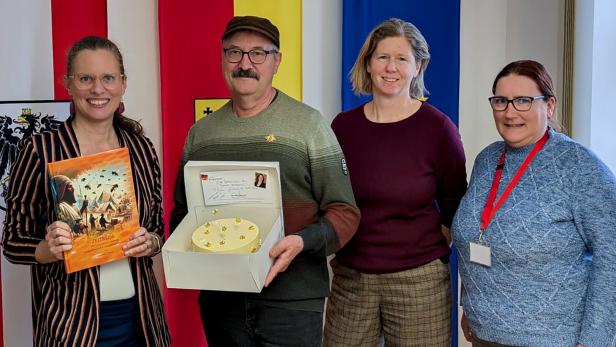 Vier Personen stehen lächelnd vor bunten Fahnen, eine Frau hält ein Buch, ein Mann eine Torte mit Karte.