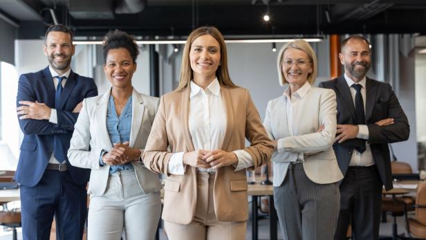 Fünf Geschäftsleute in Anzügen stehen lächelnd und selbstbewusst in einem modernen Büro zusammen.