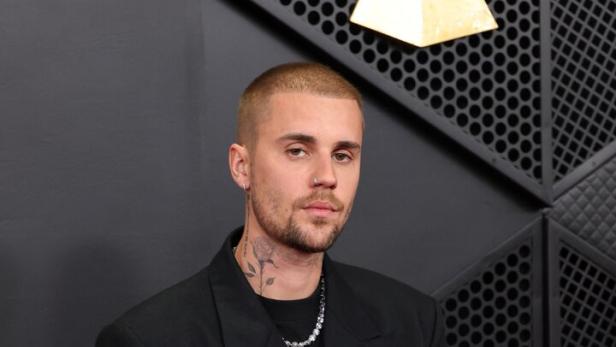 Justin Bieber trägt einen schwarzen Blazer und eine silberne Halskette vor einem dunklen Hintergrund.