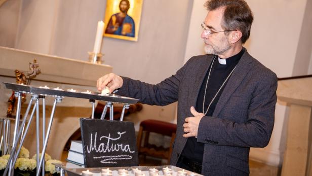 Ein Geistlicher zündet in einer Kirche eine Kerze an, im Hintergrund ein Ikonenbild und eine Tafel mit der Aufschrift „Matura“.
