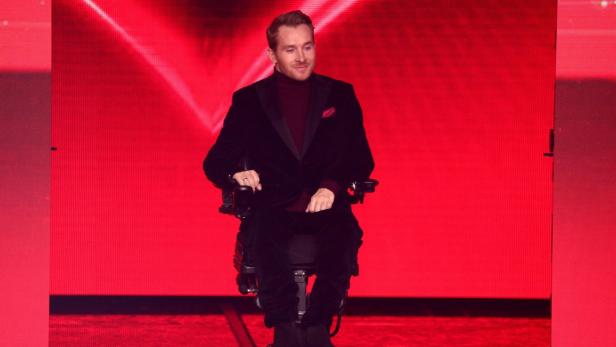 Samuel Koch im Rollstuhl vor leuchtend rotem Hintergrund, elegant gekleidet und mit leichtem Lächeln.