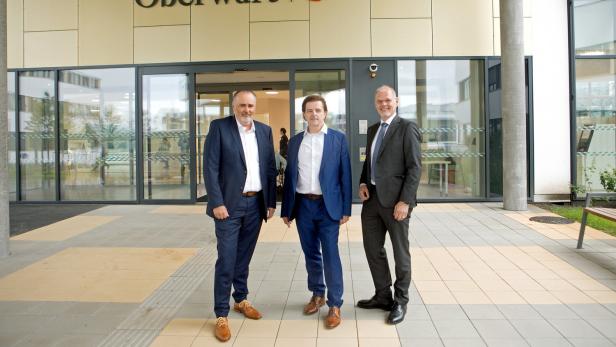 Drei Männer in Anzügen stehen vor dem Eingang der Klinik Oberwart mit Glasfassade und Klinikschriftzug.