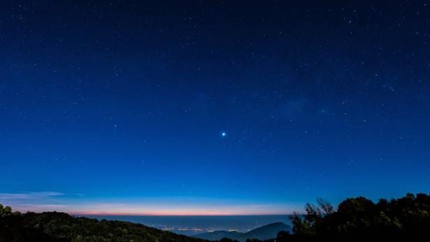 Ein heller Stern leuchtet am klaren Nachthimmel über einer bewaldeten Hügellandschaft mit fernen Lichtern am Horizont.