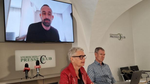 Mann und Frau an Tisch, im Hintergrund Bildschirm mit Bild eines Mannes