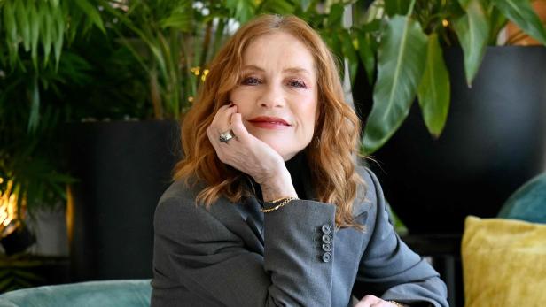 Isabelle Huppert mit rötlichem Haar und grauem Blazer sitzt an einem Tisch mit Pflanzen im Hintergrund.