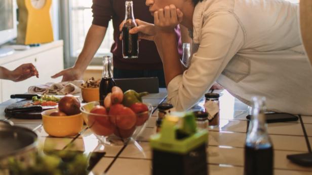 Drei junge Menschen unterhalten sich in einer Küche mit Obst und Getränken auf dem Tisch.