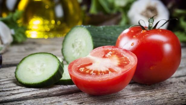Eine ganze Tomate, eine halbierte Tomate und mehrere Gurkenscheiben liegen auf einem Holztisch.