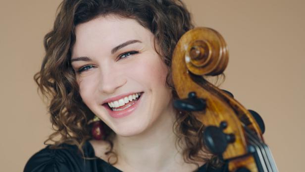 Junge Frau mit lockigen Haaren lächelt und hält ein Streichinstrument mit sichtbarem Wirbel neben ihrem Gesicht.