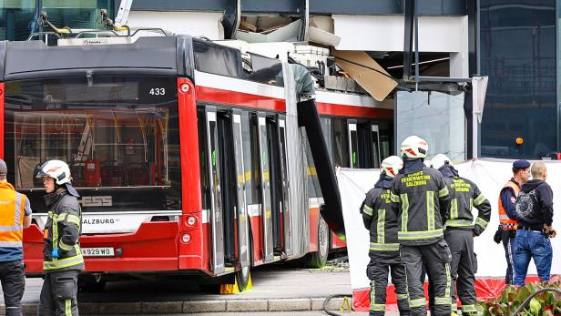 Ein roter Bus ist in ein Gebäude gefahren, Feuerwehrleute der Feuerwehr Salzburg sind vor Ort.