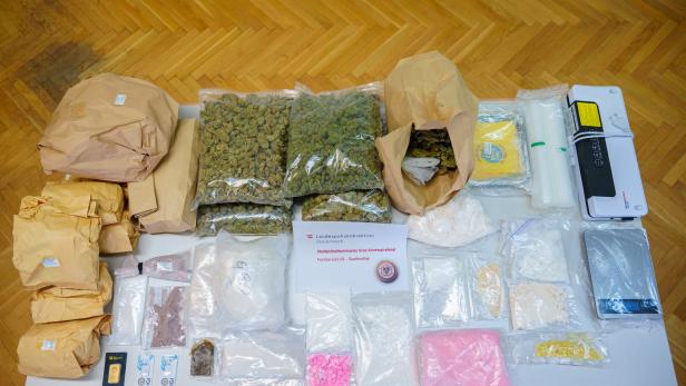 Auf einem Tisch liegen beschlagnahmte Drogen, darunter Cannabis, Pulver und Pillen, sowie eine Goldbarren und eine Waage.