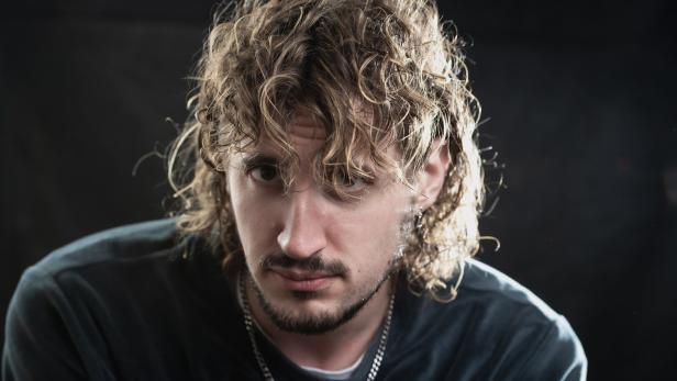 Mann mit lockigen, schulterlangen Haaren, Bart und Kette blickt ernst in die Kamera vor dunklem Hintergrund.