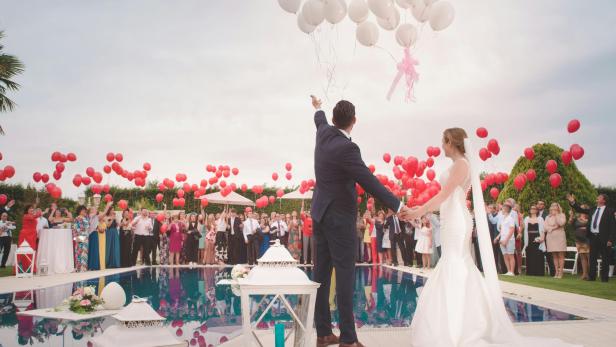 Brautpaar steht am Pool, Gäste lassen rote und weiße Luftballons steigen.