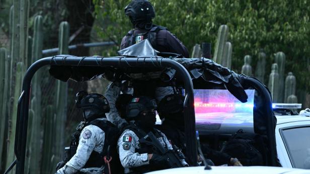 Mehrere schwer bewaffnete Einsatzkräfte in Uniform sitzen auf einem offenen Fahrzeug mit eingeschaltetem Blaulicht vor Kakteen.