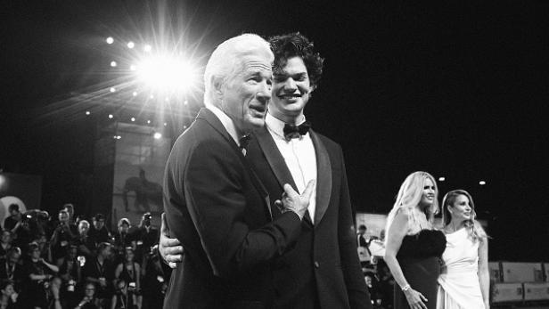 Richard Gere und sein Sohn Homer James Jigme Gere stehen gemeinsam im Anzug auf einem roten Teppich und lächeln, umgeben von weiteren Gästen.
