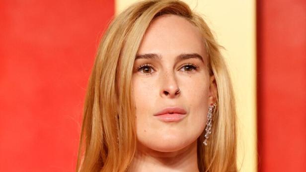 Rumer Willis mit blonden Haaren und einem rosa Kleid posiert vor einem roten Hintergrund.