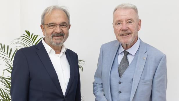 Zwei ältere Männer in Anzügen stehen nebeneinander vor einer weißen Wand mit einer Pflanze im Hintergrund.