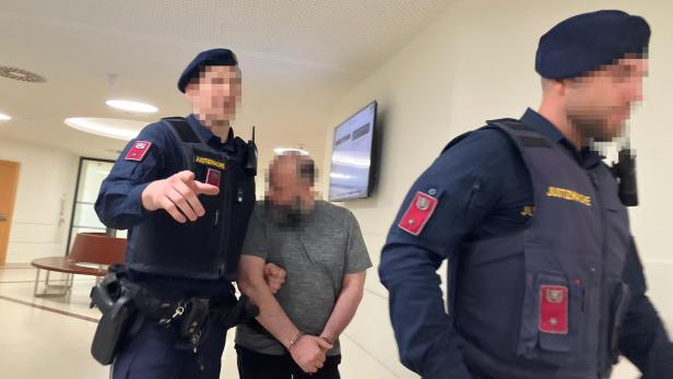 Zwei Justizwachebeamte führen einen gefesselten Mann durch einen Gerichtsflur.